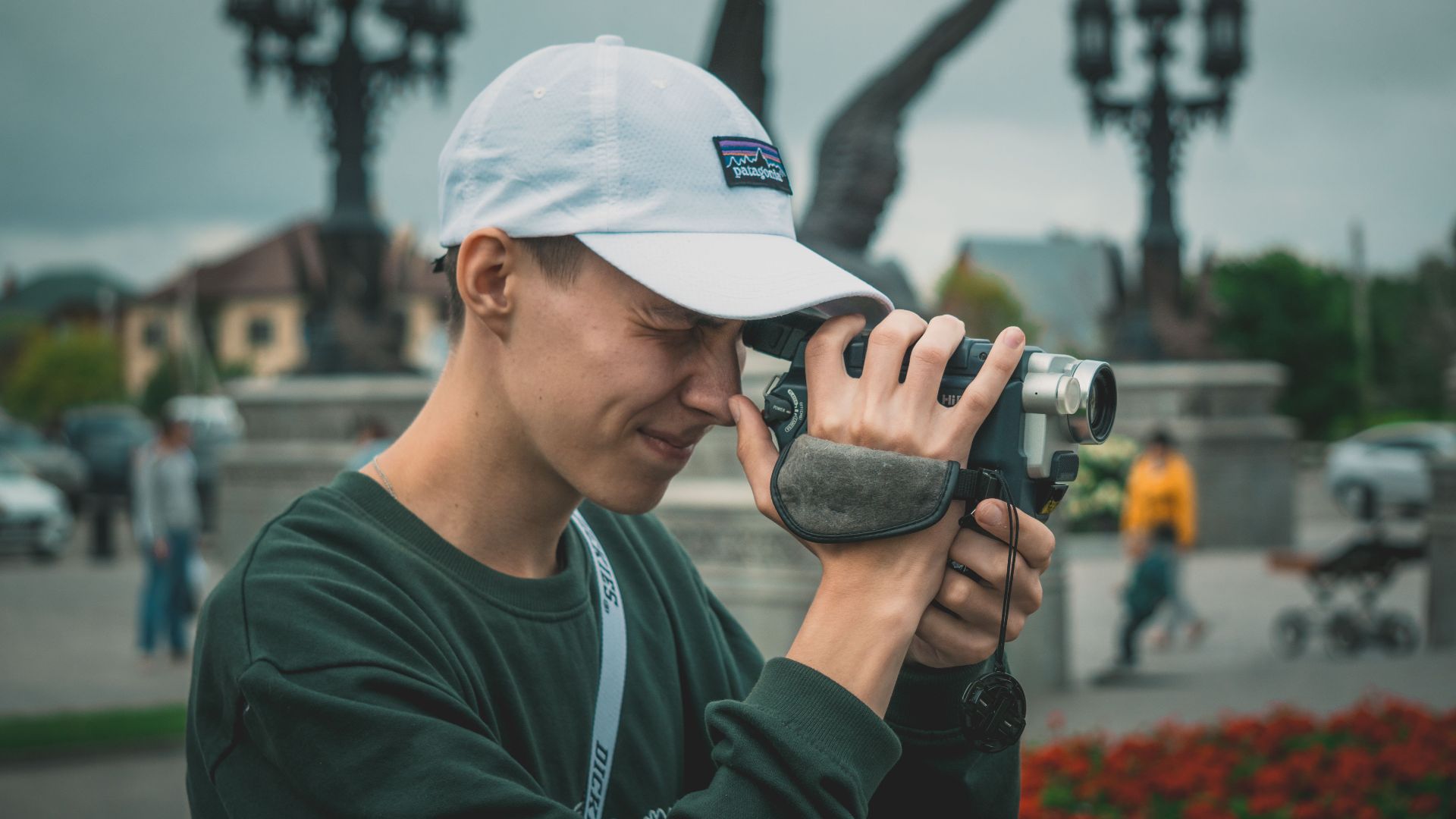 Young man concentrating while filming with a camcorder outdoors, wearing a baseball cap.