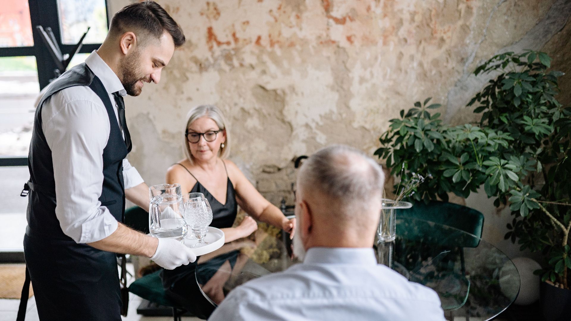 A waiter serving drinks to a senior couple in a stylish, rustic restaurant setting.