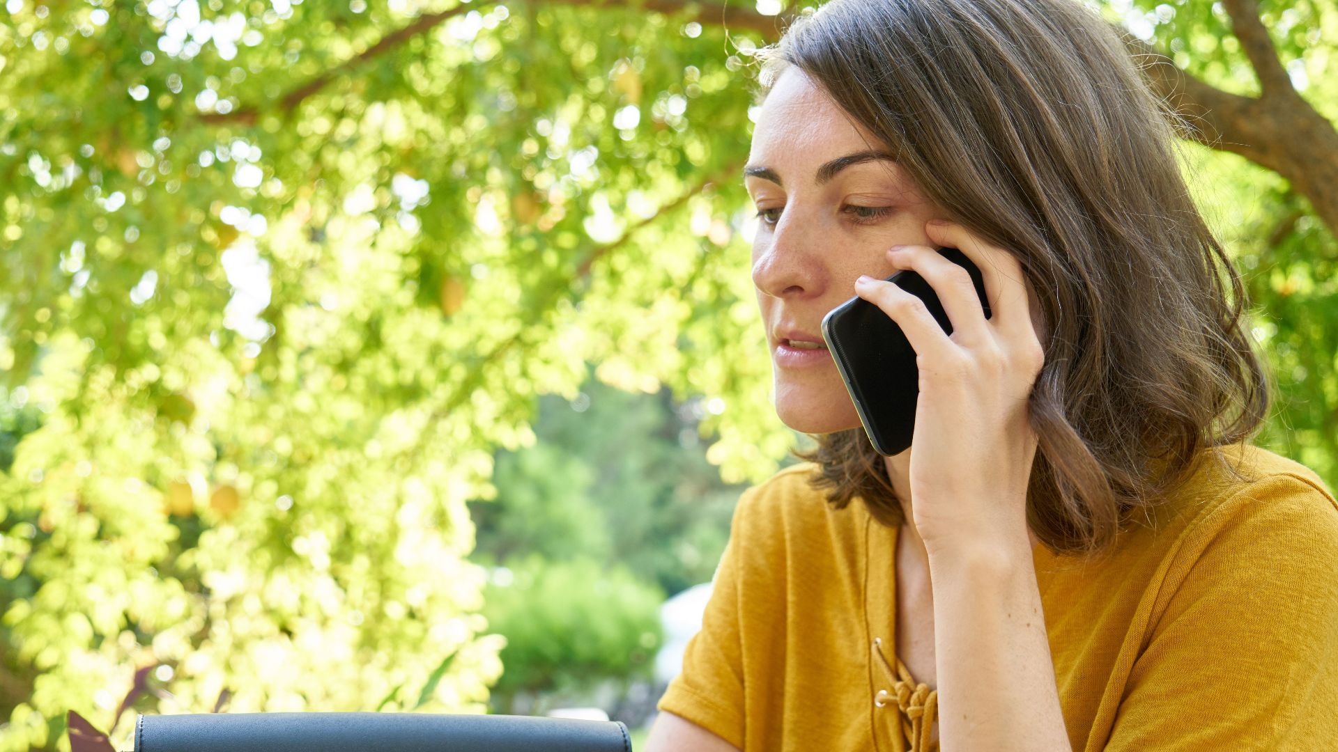 A woman in a yellow shirt makes a phone call outdoors amid lush greenery.