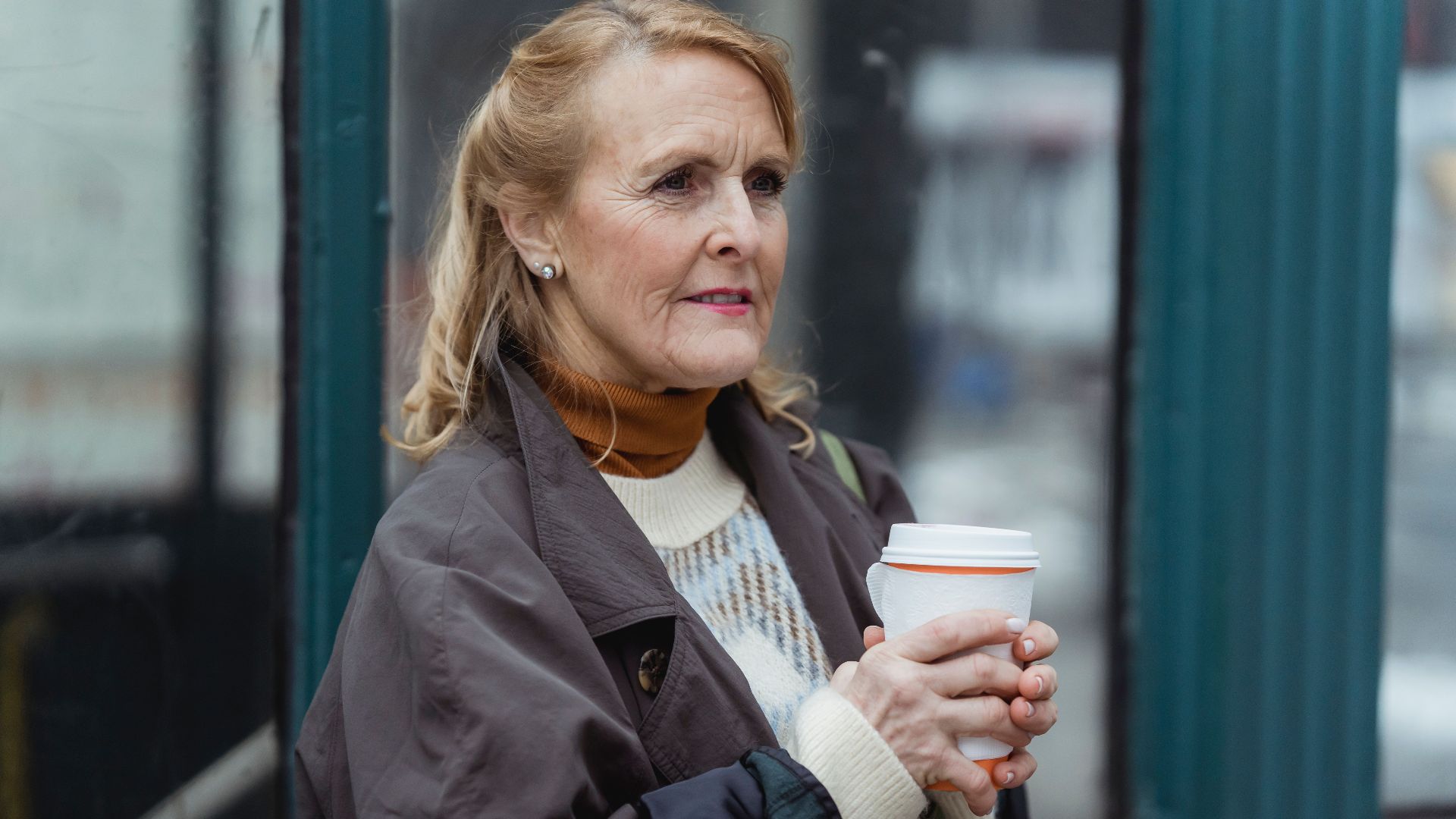 Contemplative elderly female in outerwear with hot beverage to go looking forward in town in daylight