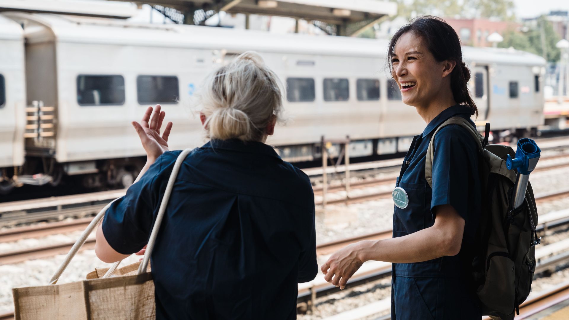 Two women laughing and talking outdoors at a busy train station during the day.