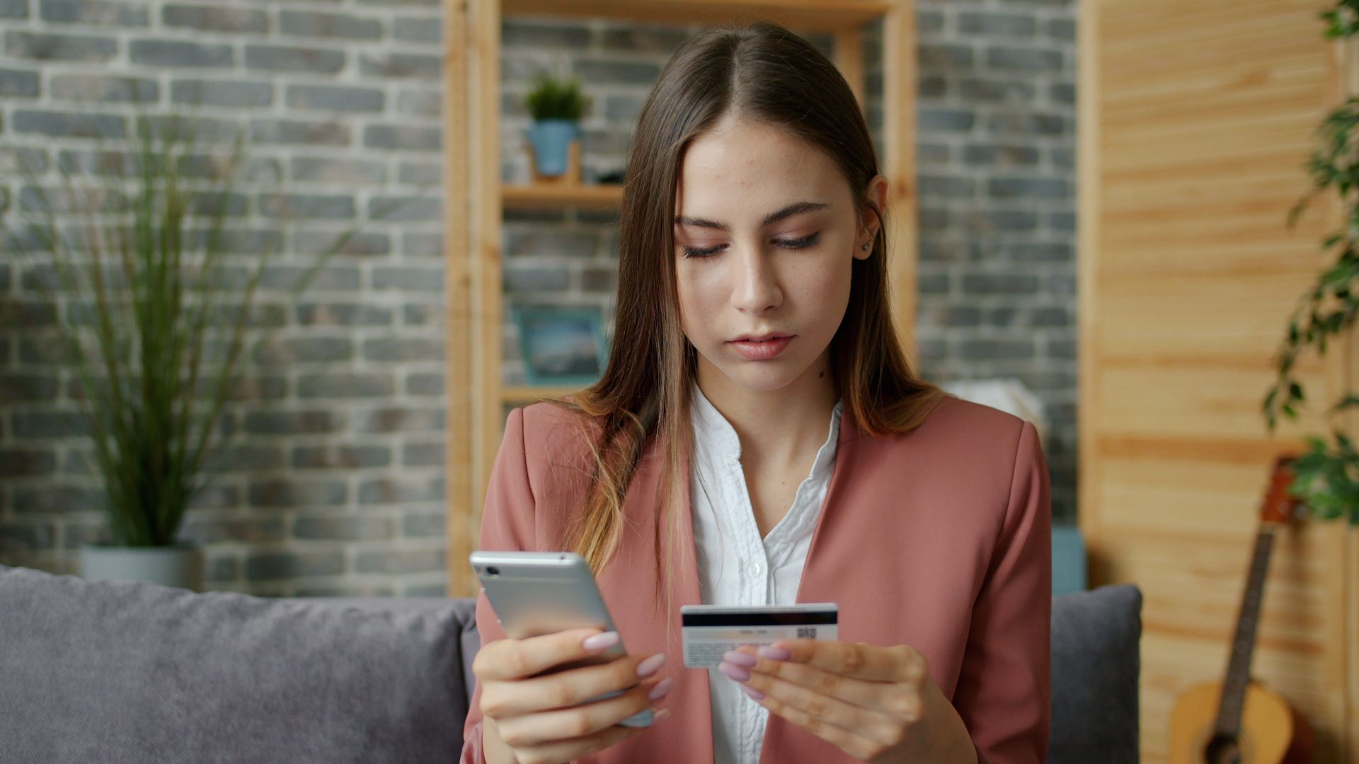 A young woman in a pink blazer shops online using a smartphone and credit card indoors.