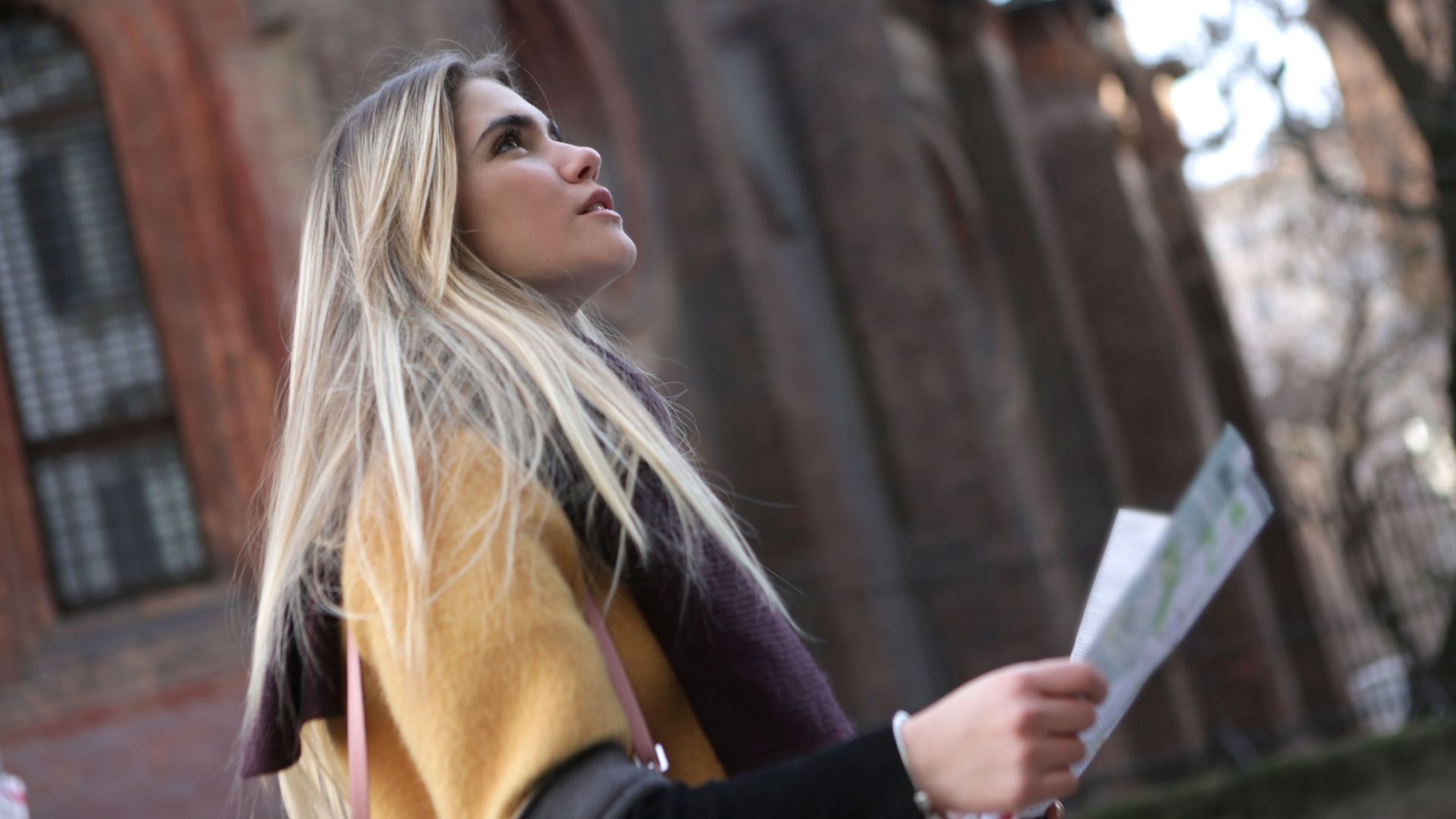 A young woman with long hair looks up while holding a map in an urban setting.