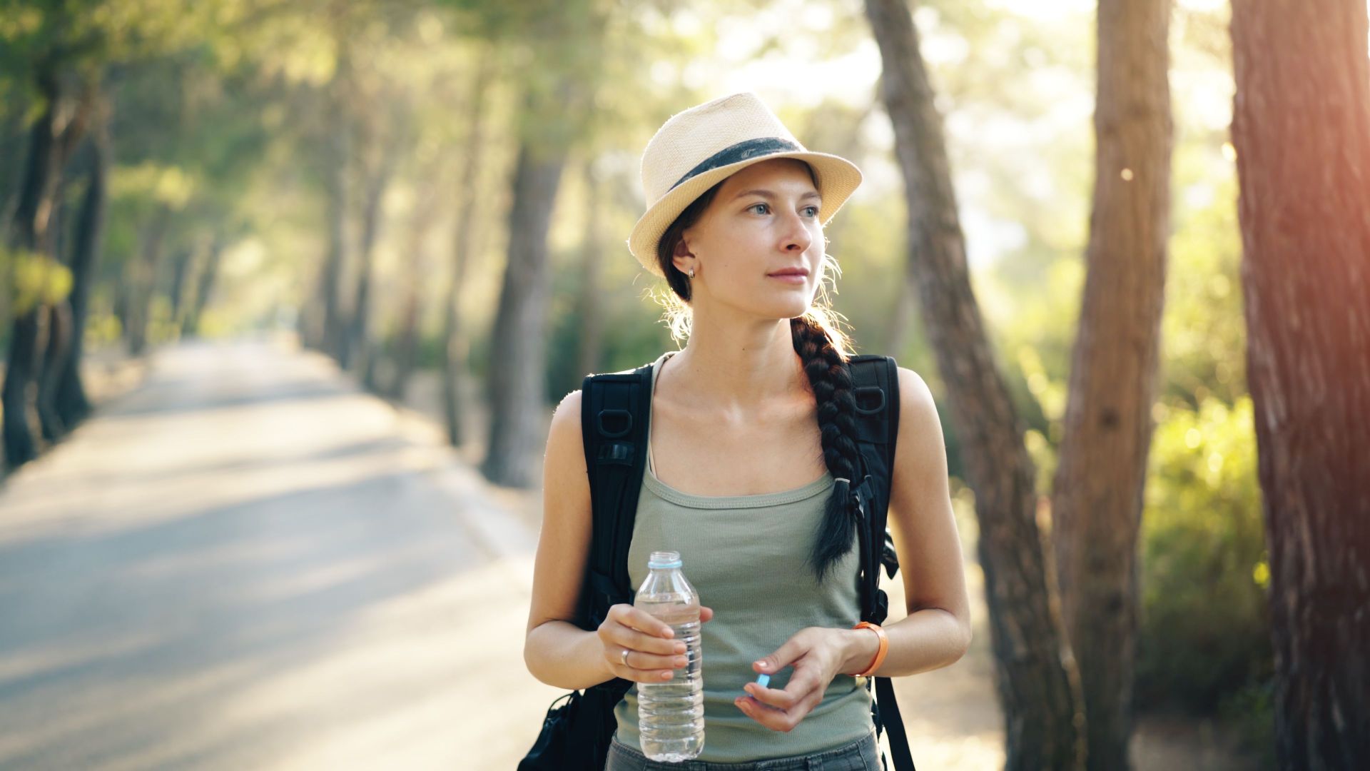 A woman with a braid and hat hiking in a sunlit forest, carrying a water bottle.