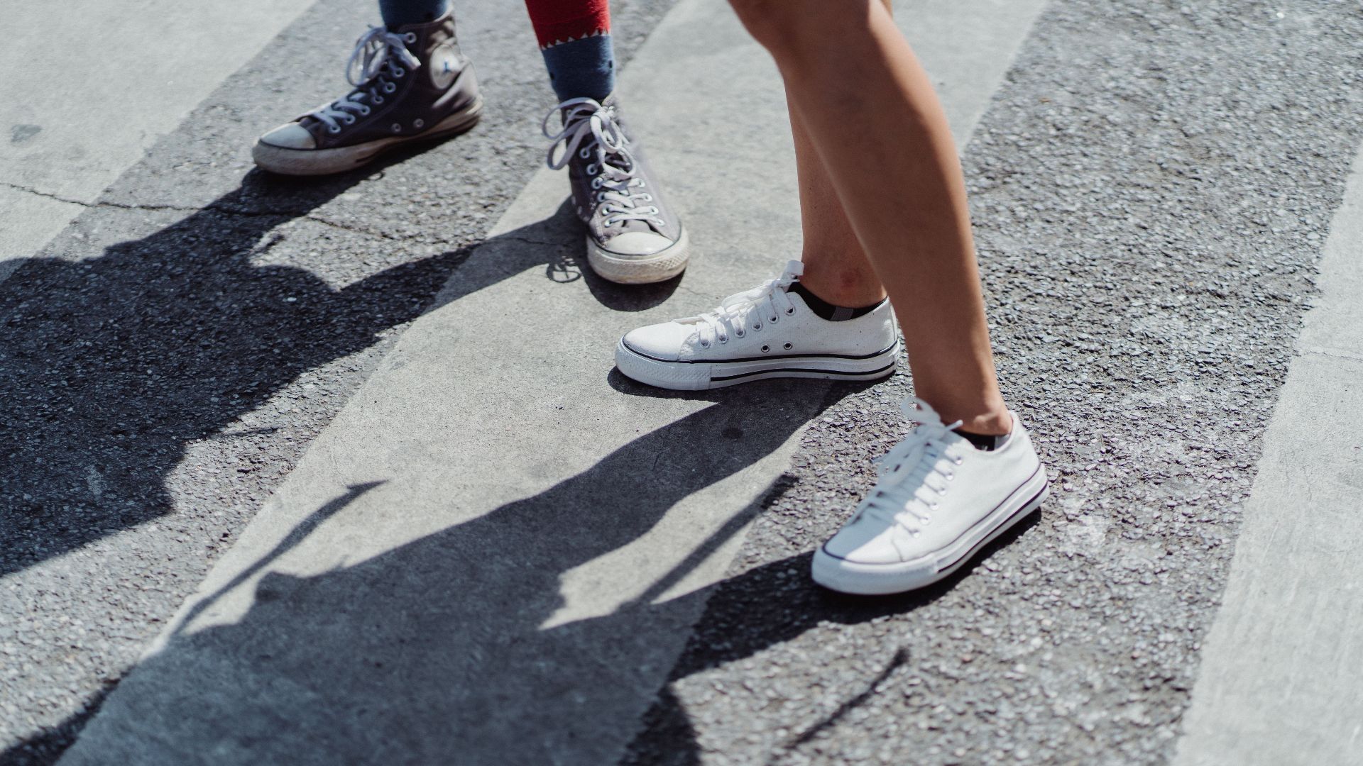 Two individuals in canvas sneakers standing on a city street crosswalk on a sunny day.