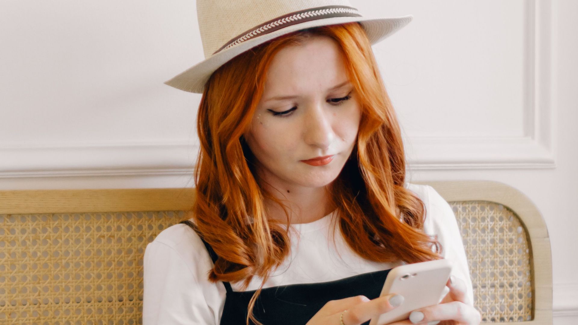 A young woman in a fedora hat texting on her smartphone while sitting with a laptop indoors.