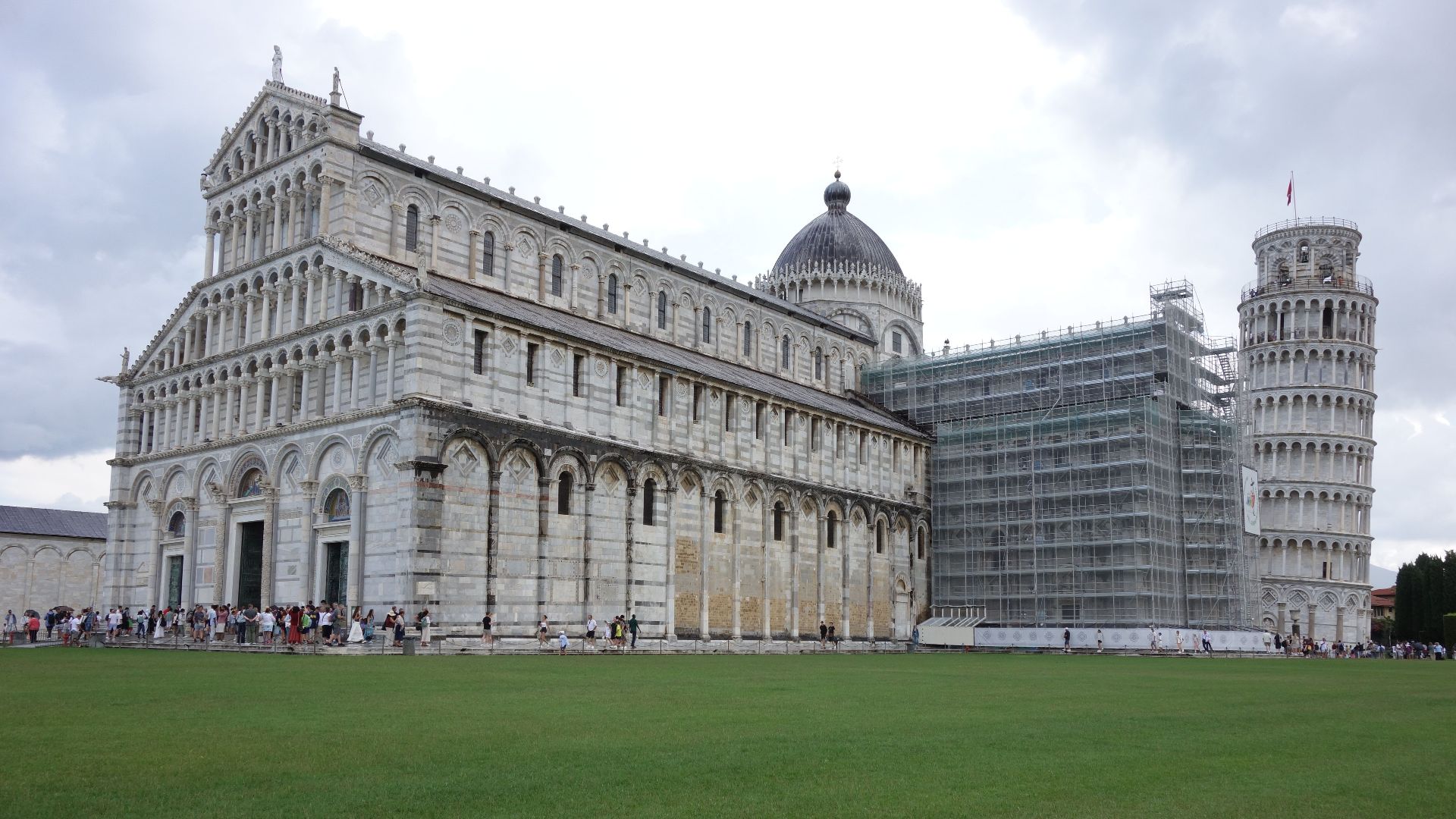 View of the Pisa Cathedral and Leaning Tower with scaffolding, under cloudy skies.