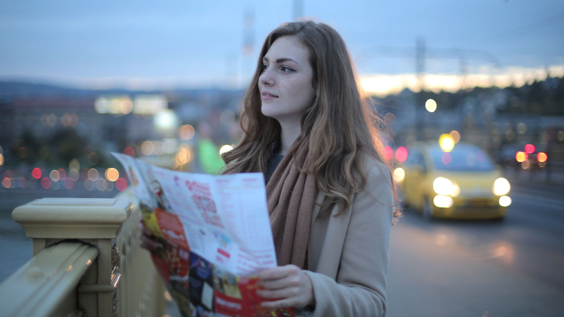 Female traveler in warm clothes standing on street with map and looking away at dusk