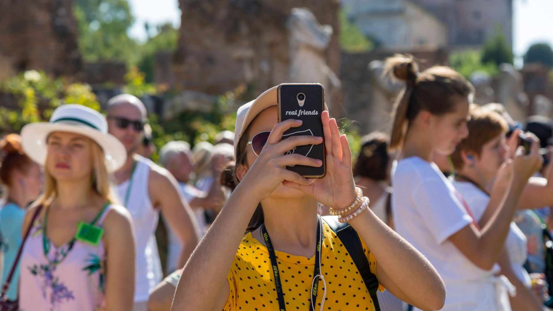 A group of tourists using smartphones to photograph historical ruins on a sunny day.