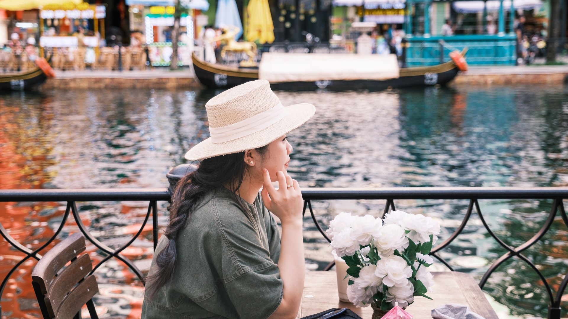 A woman in a hat sits by a riverside cafe, enjoying the view on a summer day.