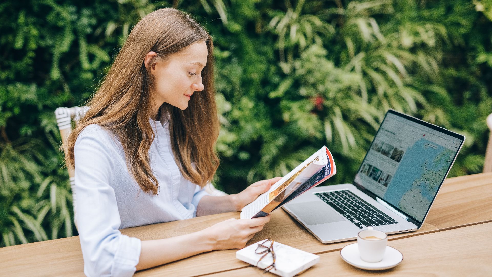 Caucasian woman reading a magazine at a wooden table with a laptop, outdoors.