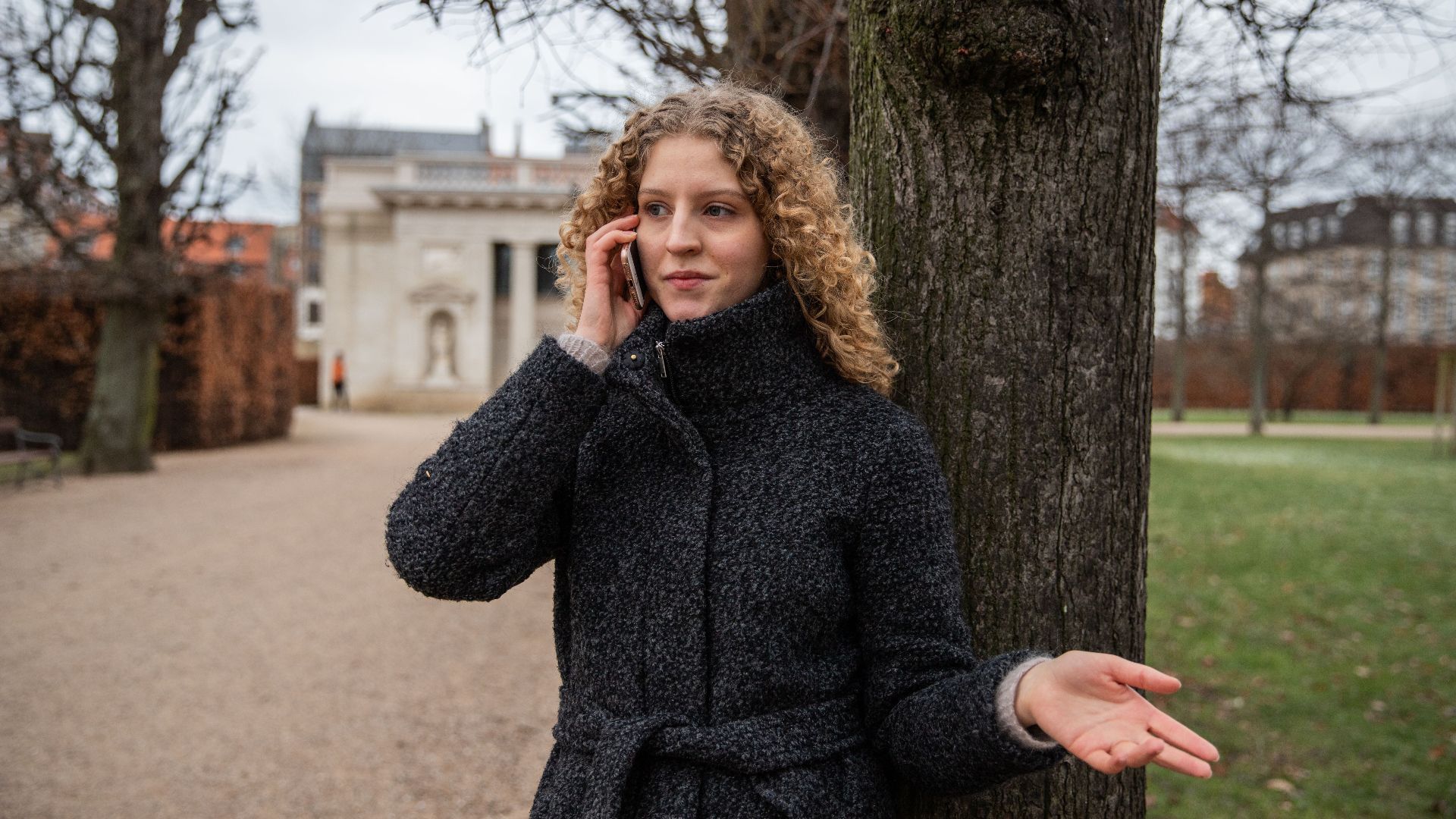 A woman in a coat talks on her phone against a tree in a Copenhagen park.