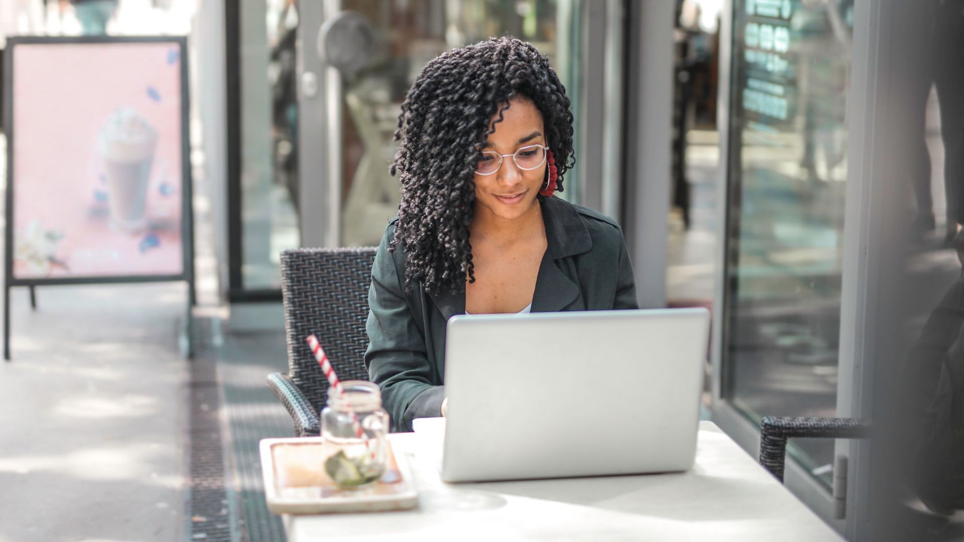 High angle of pensive African American female freelancer in glasses and casual clothes focusing on screen and interacting with netbook while sitting at table with glass of yummy drink on cafe terrace in sunny day