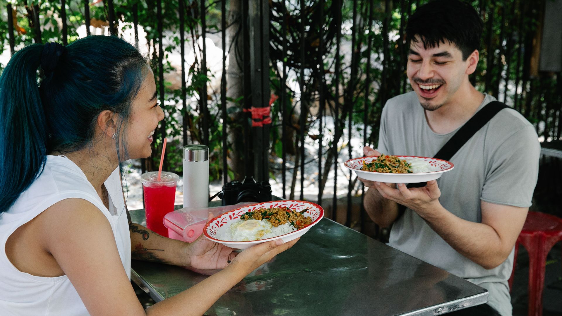 Happy multiracial couple sitting at table with plates of delicious food while having lunch together in outdoor cafeteria on summer day