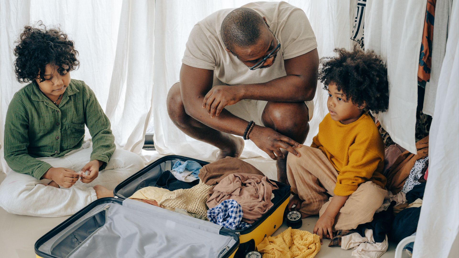 African American father in eyewear with sad kids with curly hair sitting near opened suitcase