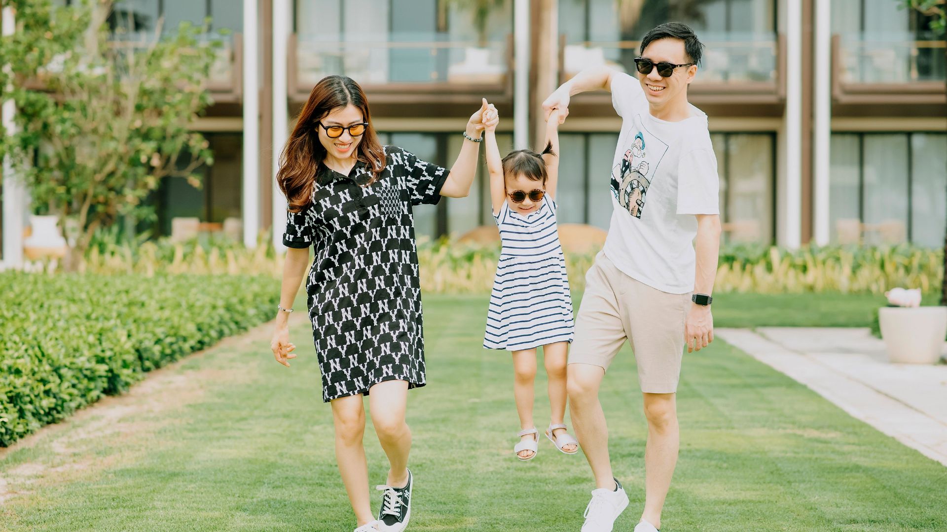 Cheerful young Asian mother and father wearing casual clothes and laughing while raising daughter on hands and standing on green lawn in front of resort territory