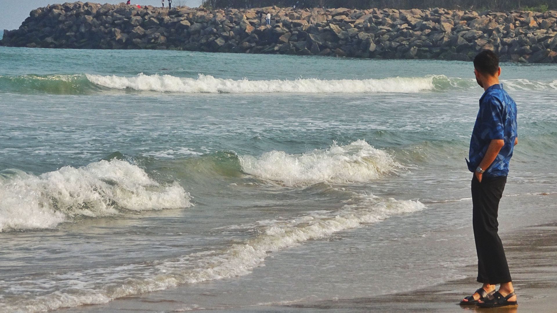 A man in casual attire reflects by the ocean waves on a peaceful beach, emphasizing solitude and contemplation.