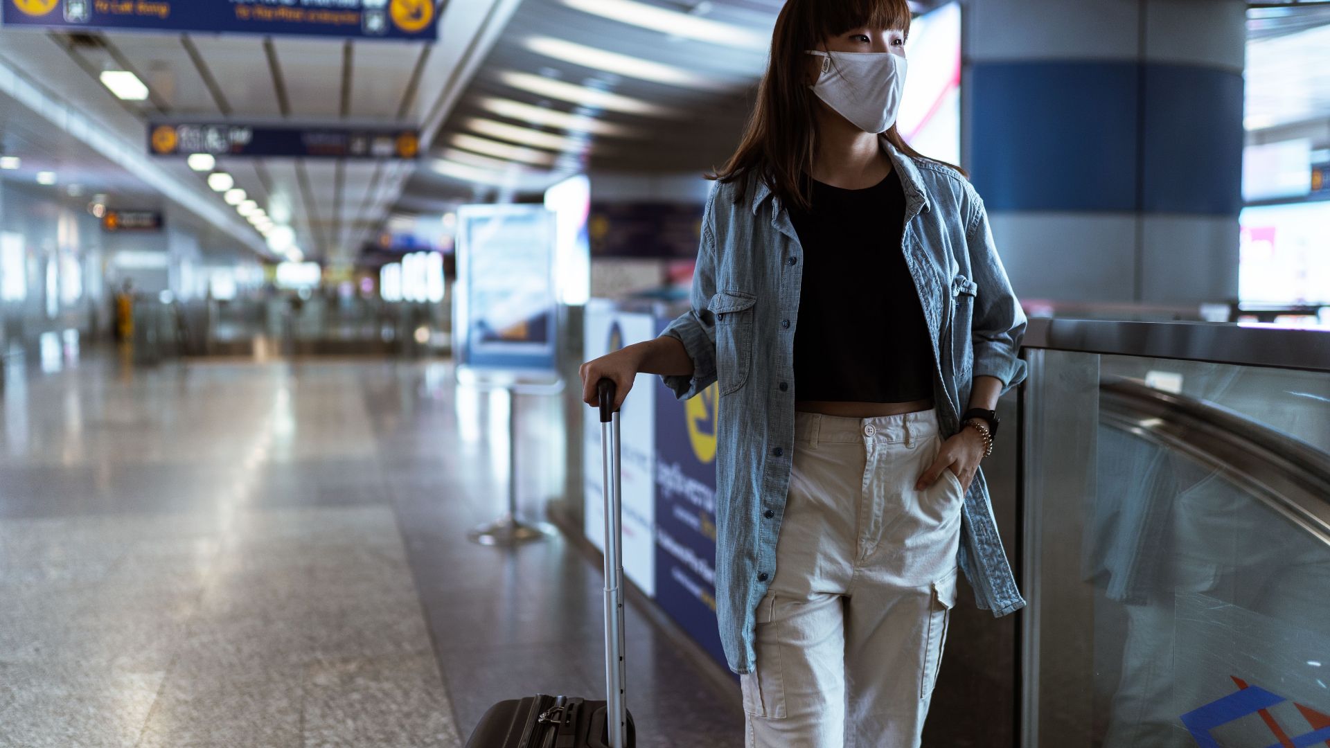 Woman wearing mask and denim jacket in airport terminal with luggage.
