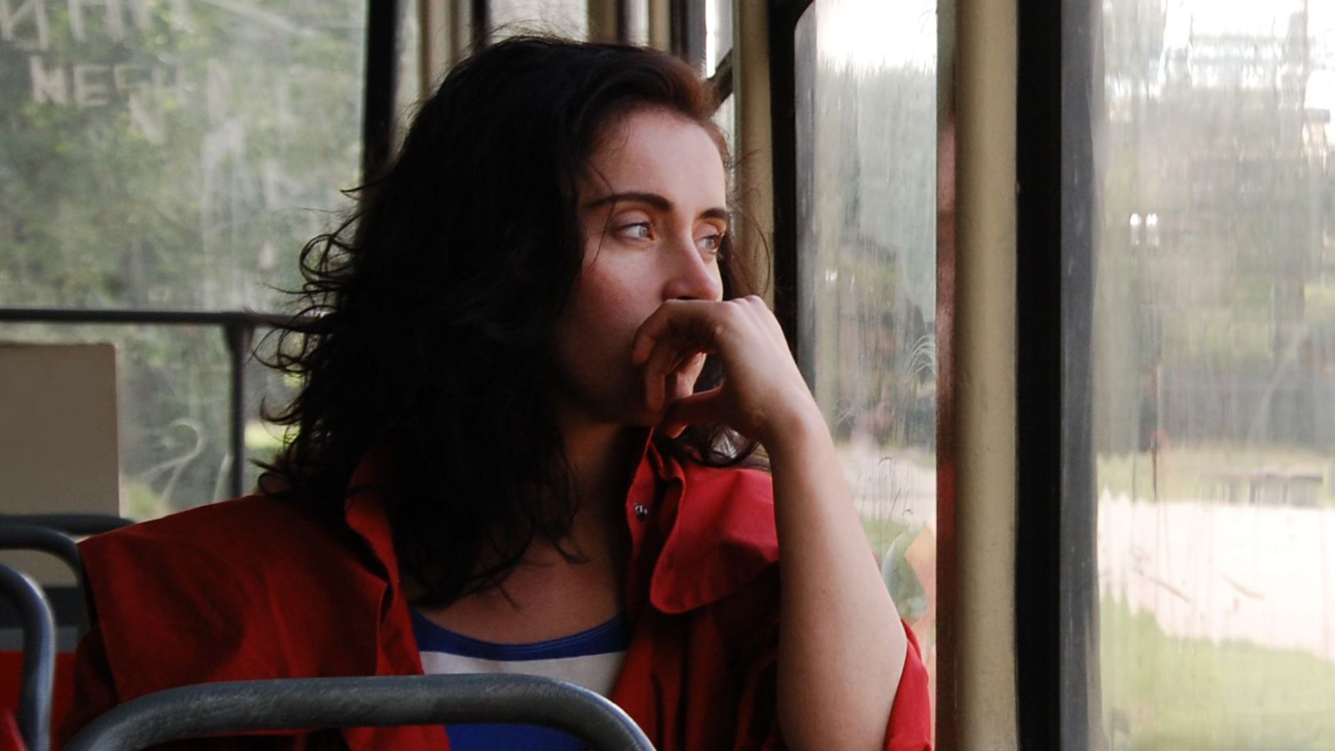 A woman in a red coat looks pensively out the window of a moving bus.
