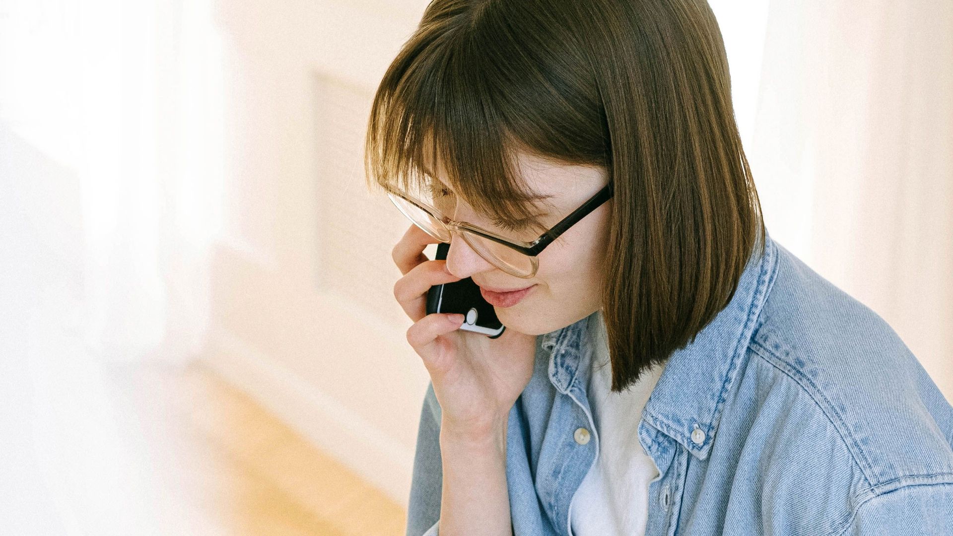 Casual young woman in denim talking on phone while enjoying a coffee indoors.