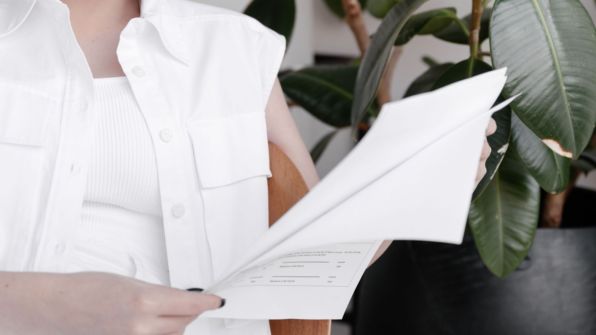 A woman with platinum hair reads a document in a stylish, plant-filled interior.