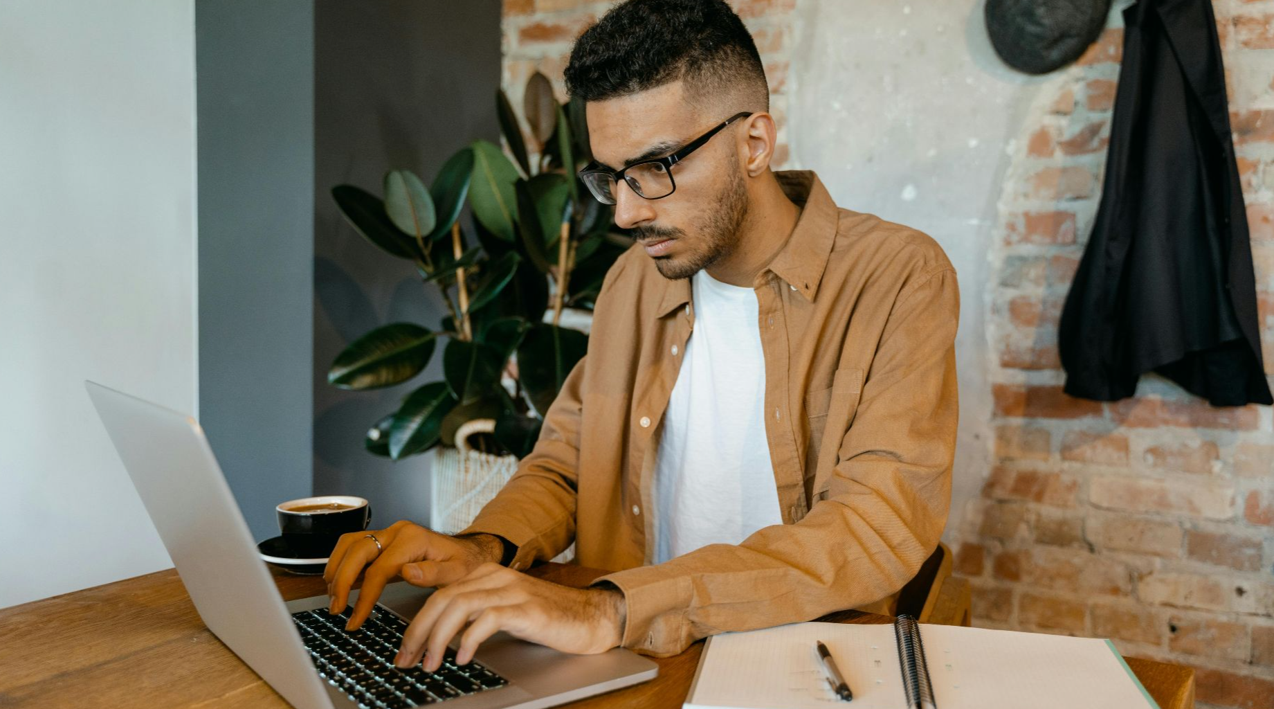 Man in casual office setting typing on laptop with coffee and notebook on desk.