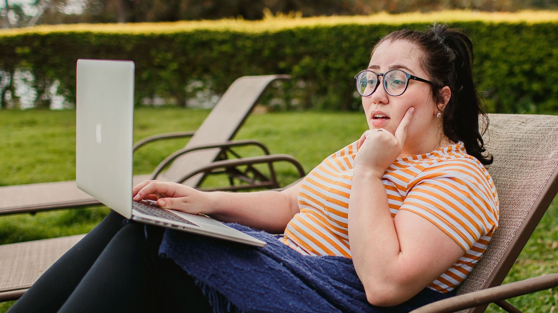 Woman relaxing on lounge chair with a laptop in a lush green garden, enjoying a sunny day.