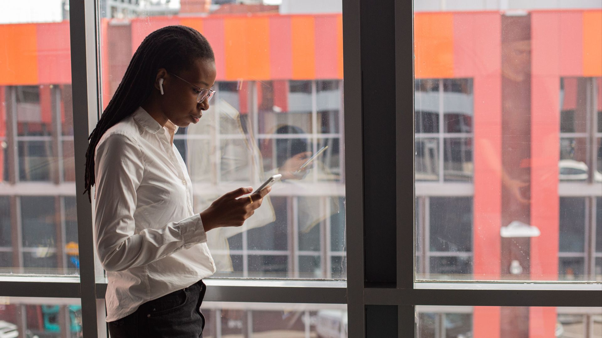 African American woman focused on her smartphone by a large office window, urban background.