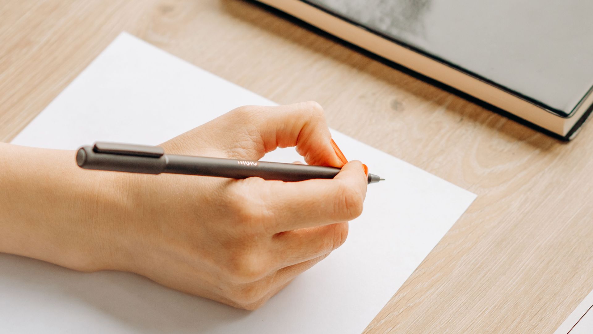 Close-up of hands writing on paper with a pen, sitting at a wooden desk indoors.