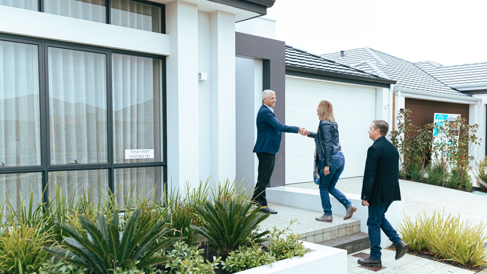 A realtor shakes hands with potential buyers outside a modern model home.