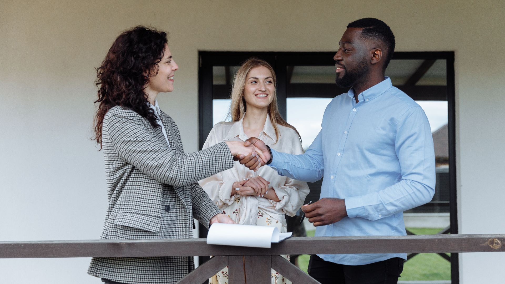 Three adults sealing a real estate deal with a handshake on a porch.