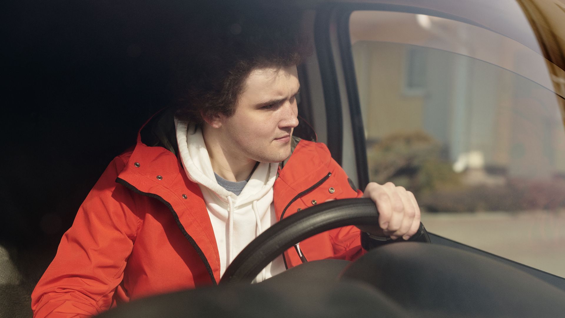 A young man in a red jacket steering a car with focus, enjoying a peaceful drive.