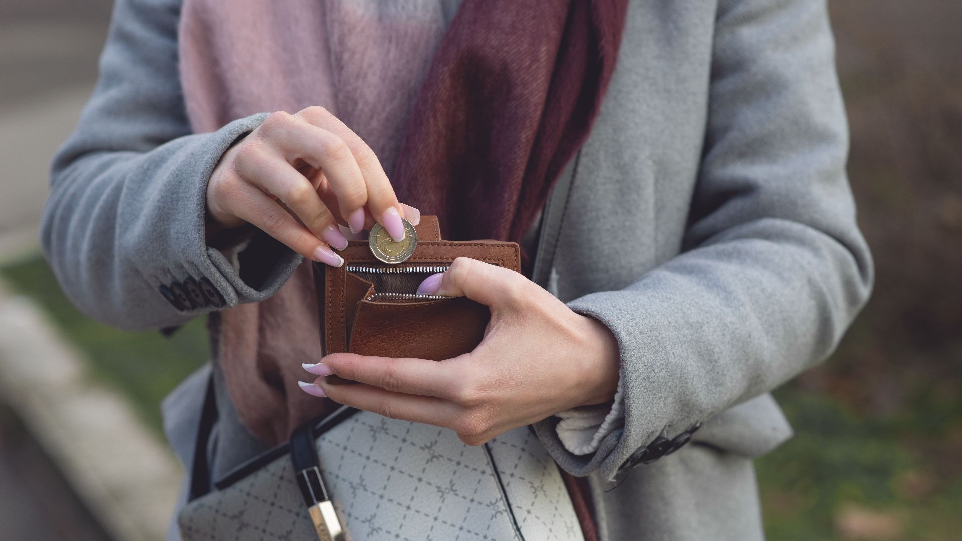 Close-up of a woman placing a coin into a wallet while outdoors, wearing a coat and scarf.