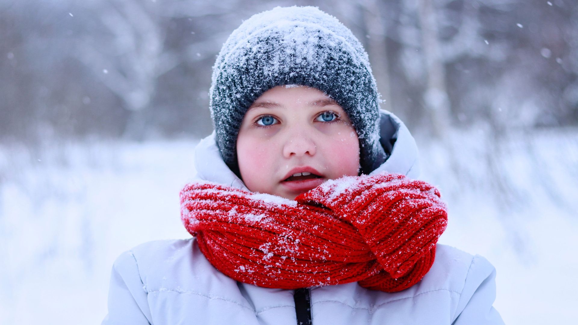 Child in winter clothing with red scarf, snow falling around.