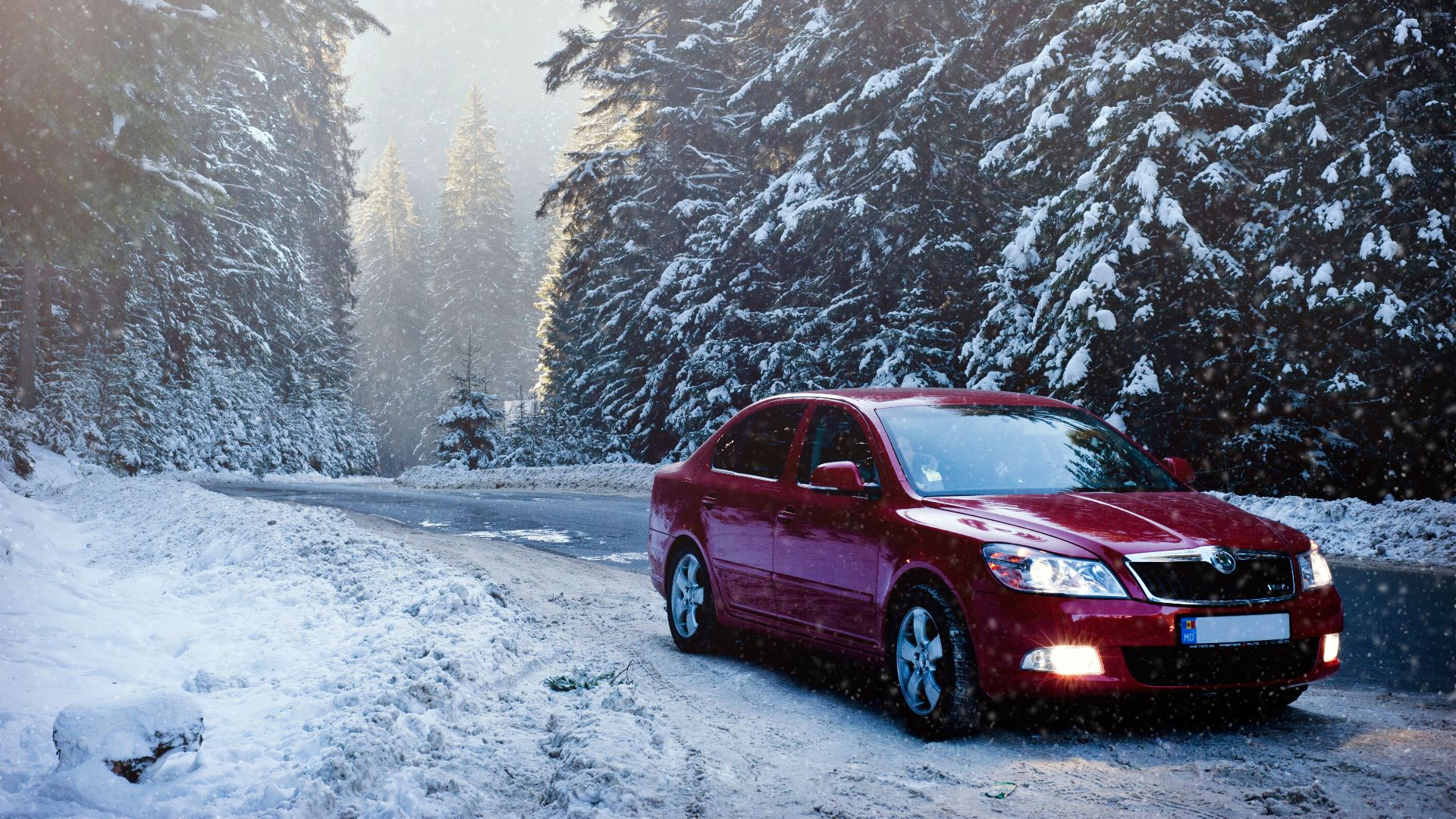 A red car drives along a snowy forest road in winter, surrounded by frosty trees.