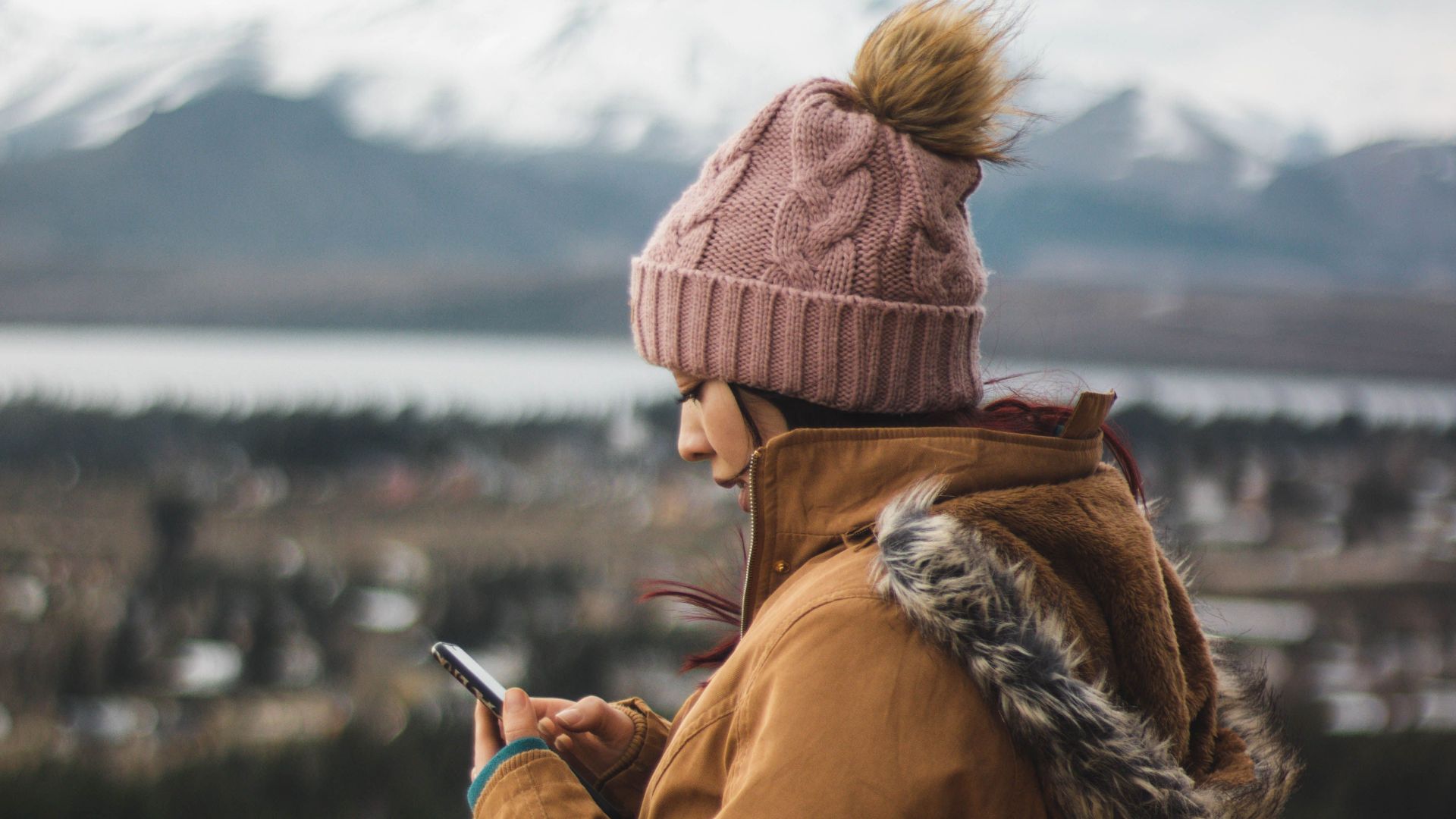 Woman wearing beanie and jacket using a smartphone outdoors with snow-capped mountains in the background.