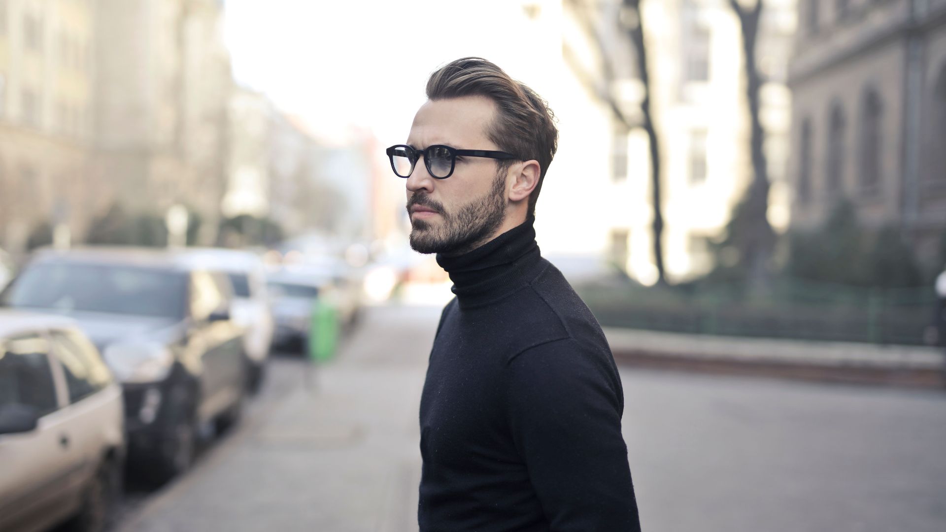 A fashionable man in a black top and glasses poses in an urban street with parked cars.