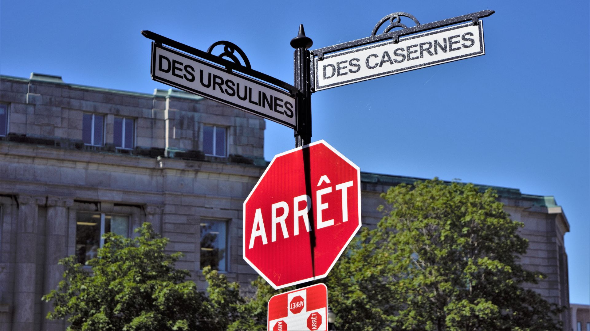 Bilingual street signs with stop sign against a clear blue sky in a city setting, featuring historical street names.