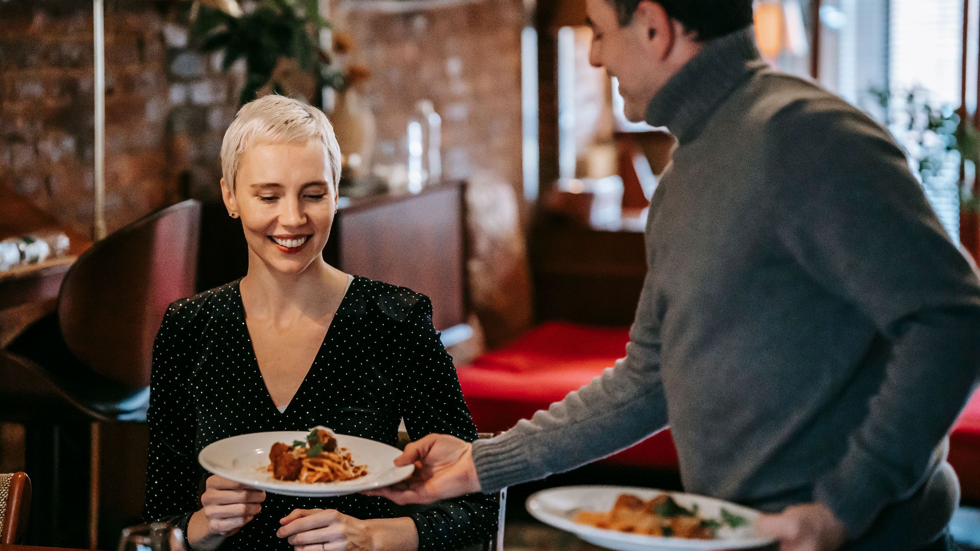 Positive adult couple having dinner in bright room near table while man serving plates with tasty pasta with meatballs near chairs in daylight