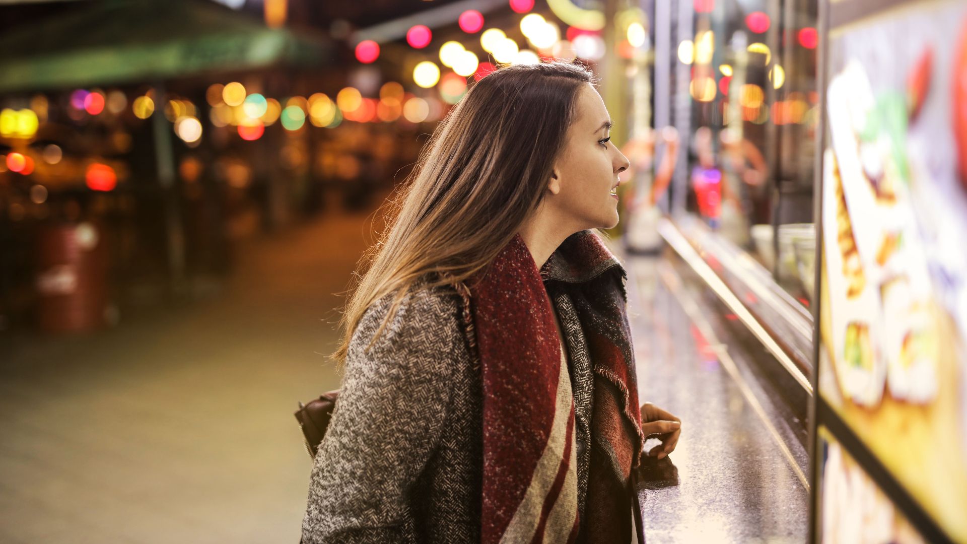 A young woman in a coat admires a shop display under vibrant city lights and bokeh effects at night.