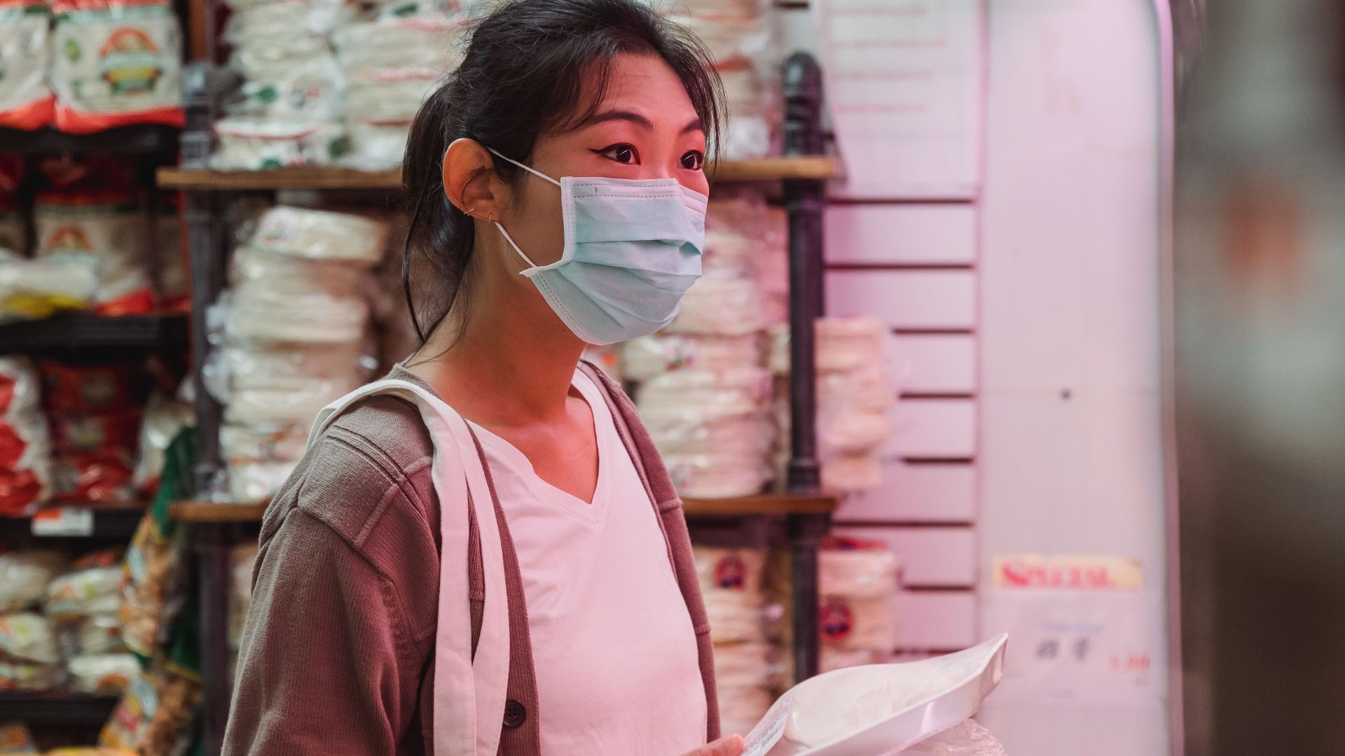 Masked Asian woman shopping in an indoor market, examining products with a thoughtful expression.