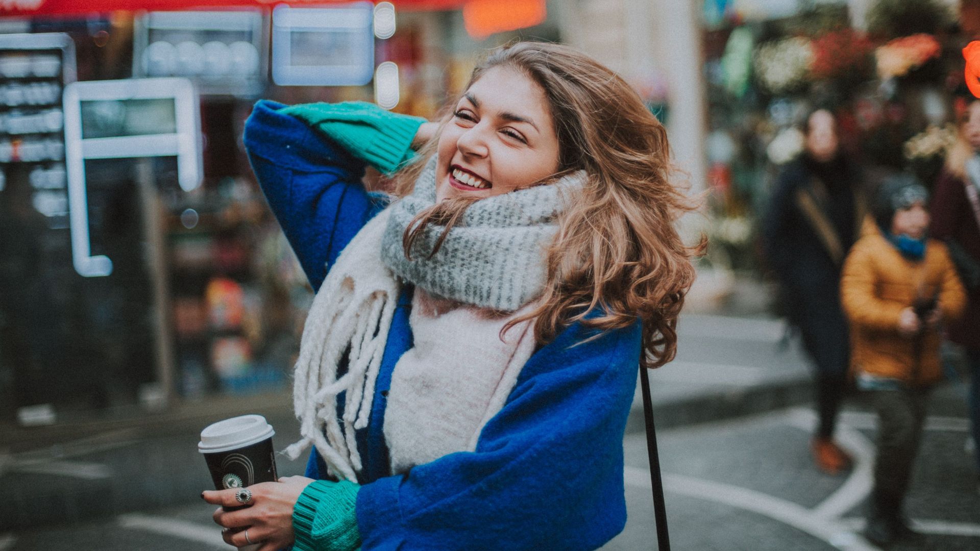 A joyful woman in a warm outfit holds coffee on a lively city street filled with people and shops.