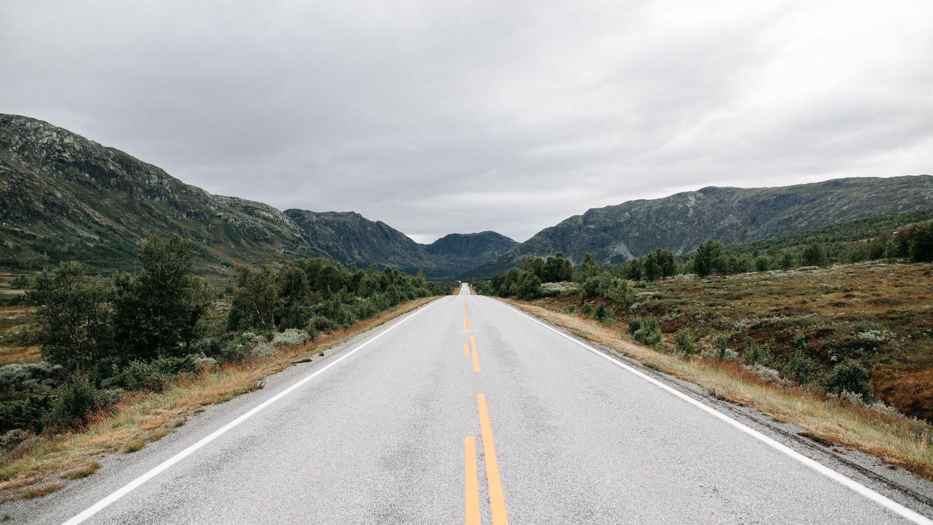An empty road stretches through a mountainous countryside landscape under an overcast sky.