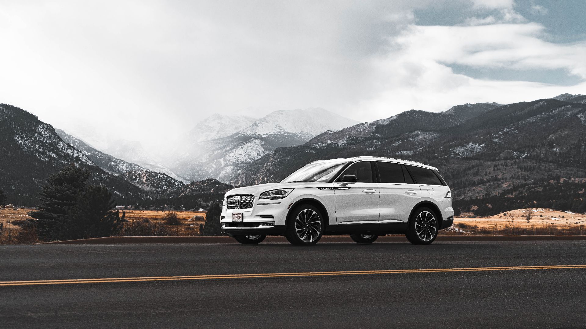 White SUV on a scenic Colorado mountain road with dramatic skies and snowy peaks.