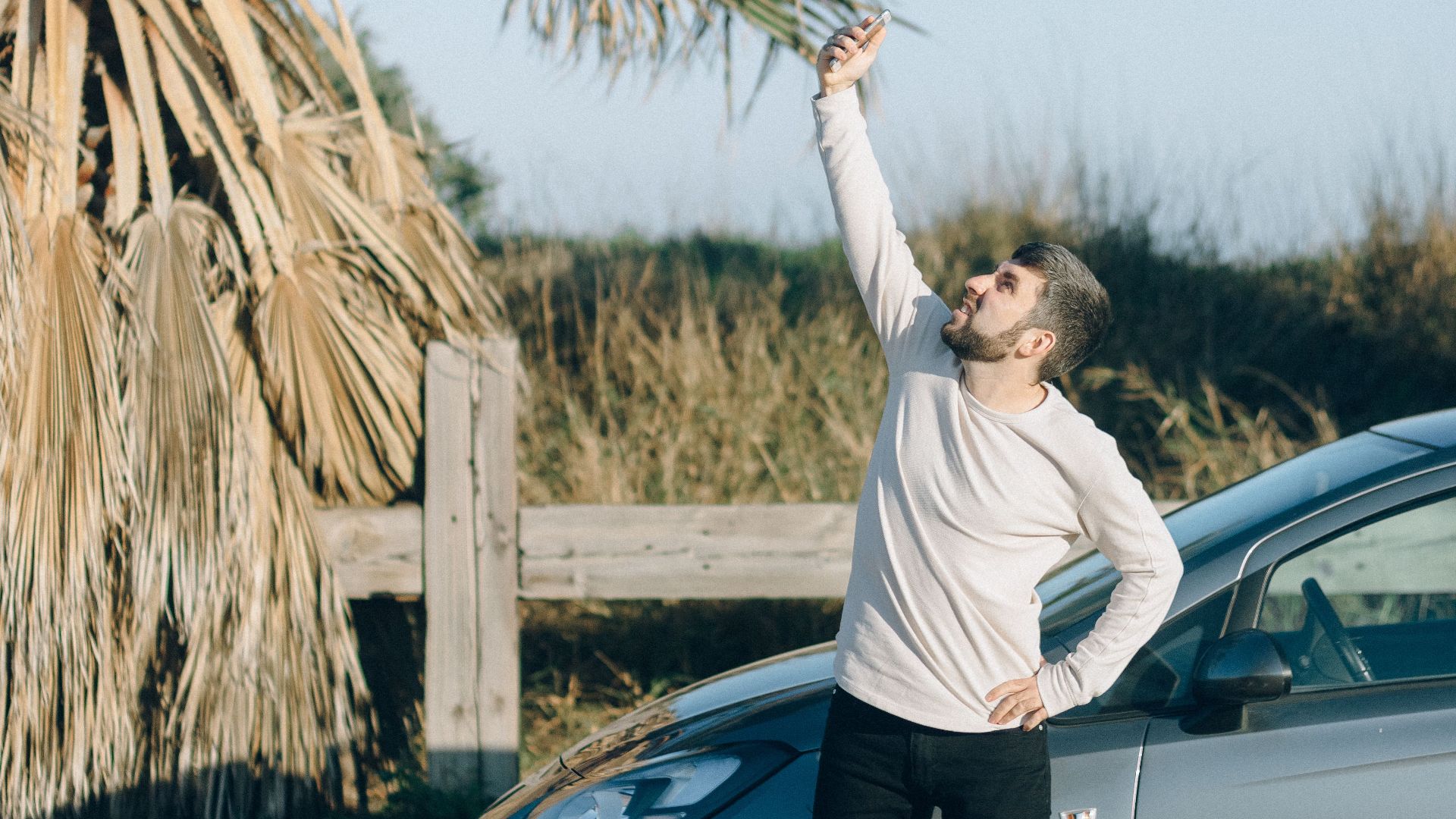 Man standing by car taking a selfie under a clear sky with palm trees nearby.