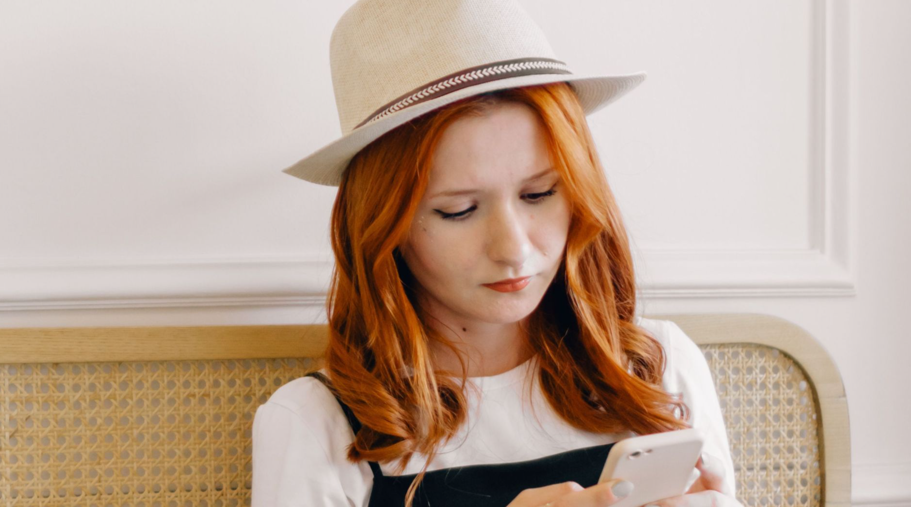 A young woman in a fedora hat texting on her smartphone while sitting with a laptop indoors.