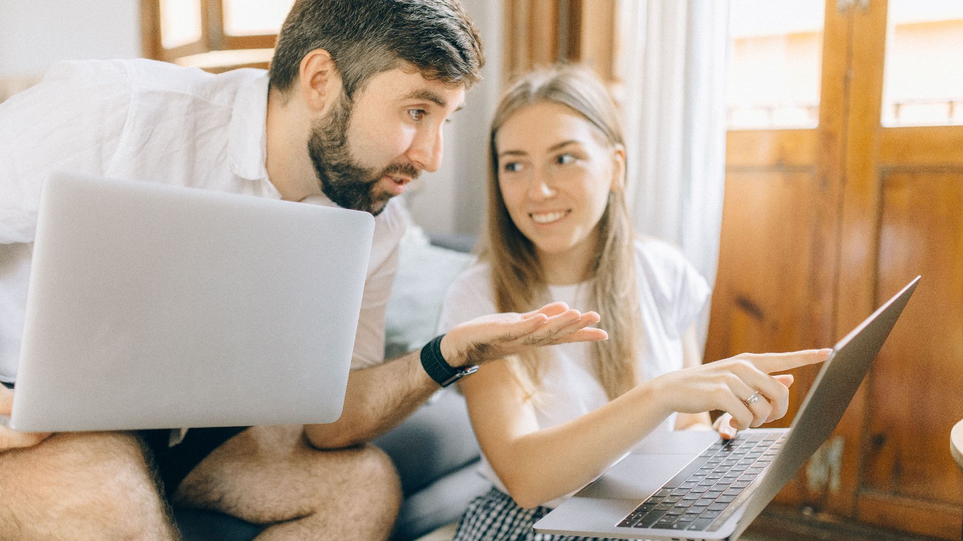 Young couple sitting together, discussing something on their laptops in a cozy room.
