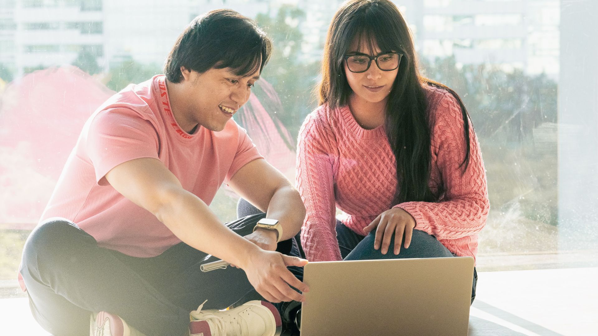 Two young adults in casual attire working together on a laptop by a window.