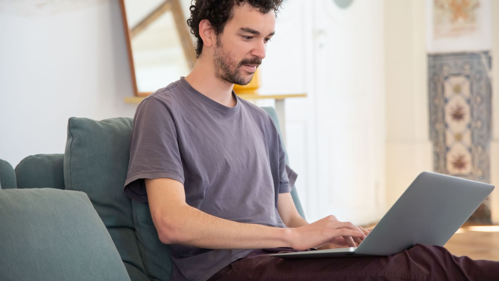 A man with curly hair typing on a laptop while sitting on a comfortable couch at home.