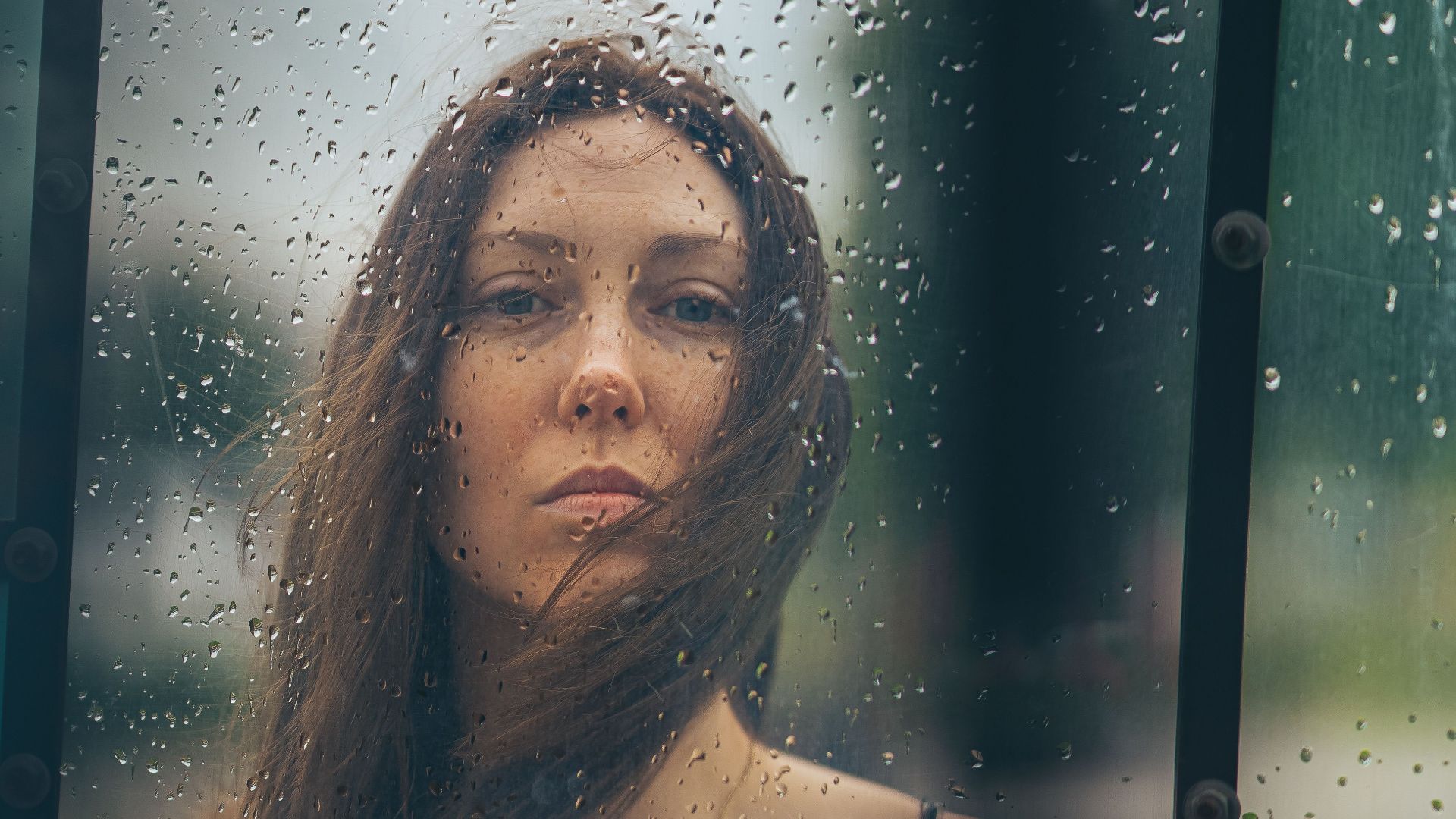 A woman in a floral dress gazes through rain-soaked glass, creating a contemplative mood.